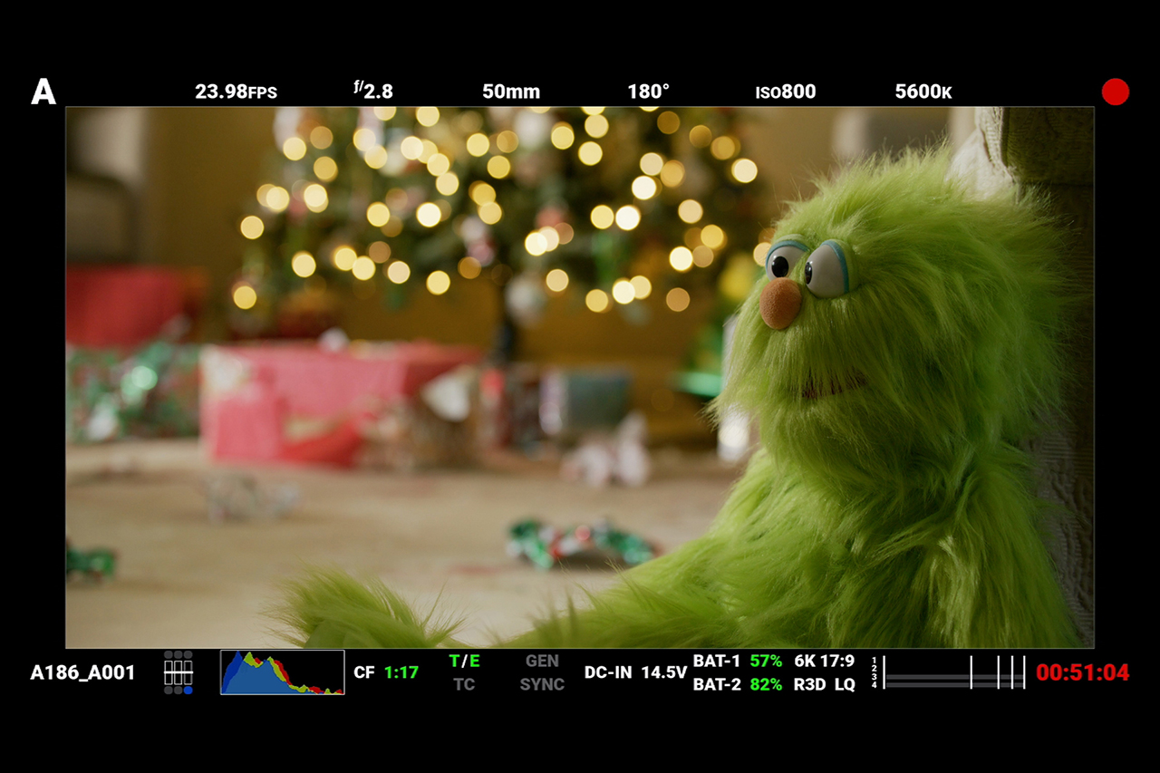 Red Digital Cinema camera monitor screenshot behind the scenes of Christmas short film, green puppet sitting on floor with Christmas decor in background..