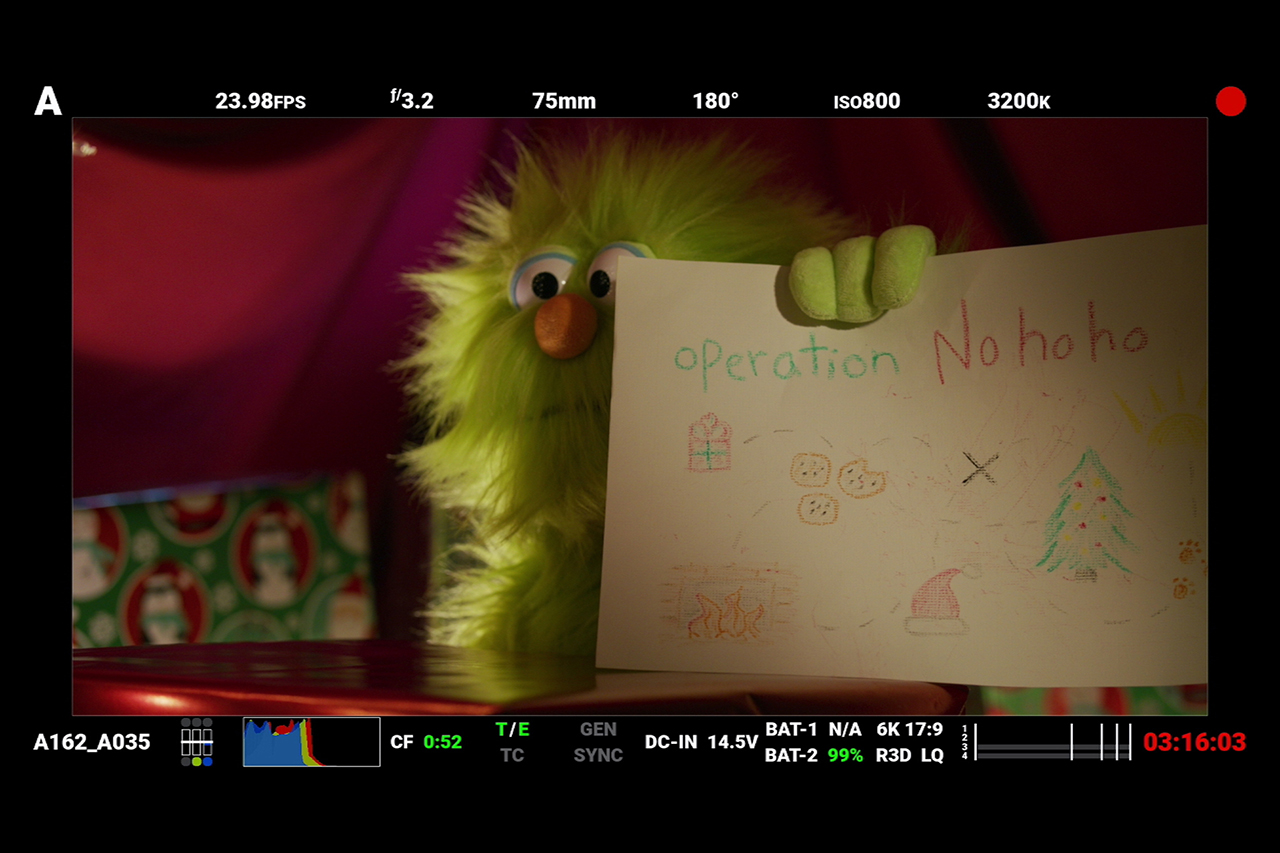 Red Digital Cinema camera monitor screenshot behind the scenes of Christmas short film, green monster puppet holding up plan to win against Santa.